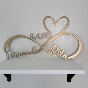 May include: Gold metal infinity symbol with a heart in the center. The names "Fernando" and "Rita" are inscribed on the symbol, along with the date "8.4.1973".