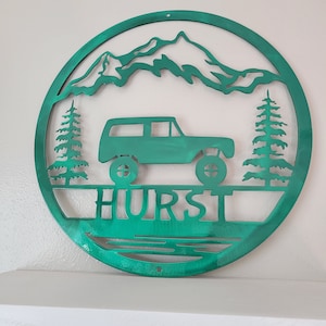 May include: A teal metal wall art piece featuring a mountain range, a vehicle, and two pine trees within a circular frame. The word "HURST" is displayed below the vehicle. The artwork is designed to be mounted on a wall.