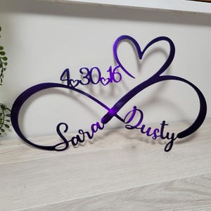 May include: A purple infinity symbol wall sign featuring a heart shape at the top, the names Sara and Dusty in cursive script, and the date 4.30.16 with small heart accents integrated into the design, crafted from a smooth, reflective material.