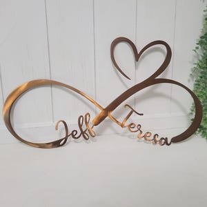 May include: A copper-colored metal wall sign featuring an infinity symbol intertwined with a heart shape. The names Jeff and Teresa are cut out in a cursive script font along the lower loops of the infinity symbol, creating a continuous design.