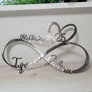 May include: A metal infinity sign with the names "Tyrone" and "Calina" in cursive script. The sign includes the date "02.02.2001" and the number "25" within a heart shape, symbolizing a wedding anniversary.