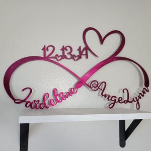 May include: A magenta metal infinity sign with a heart, dates, and names. The sign is mounted on a white shelf with black brackets. The names "Zardelaine" and "AngeLynn" are written in cursive.