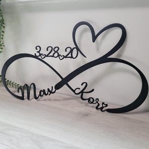 May include: Black metal infinity sign with a heart, the date 3.28.20, and the names Max and Kore. The sign is on a light-colored wooden surface with greenery in the background. A decorative piece for a wedding or anniversary.