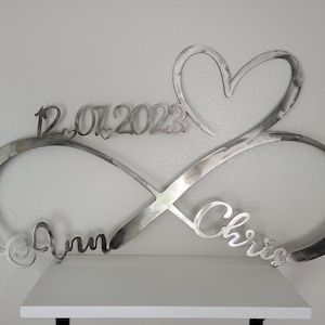 May include: Silver metal infinity symbol with a heart at the top and the names "Ann" and "Chris" on either side. The date "12.07.2023" is inscribed above the heart.