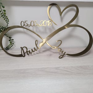 May include: A gold-colored metal wall decoration featuring an infinity symbol intertwined with a heart shape. The piece includes the names Paul and Joy in cursive script and the date 05.05.2018 with small heart accents between the numbers.