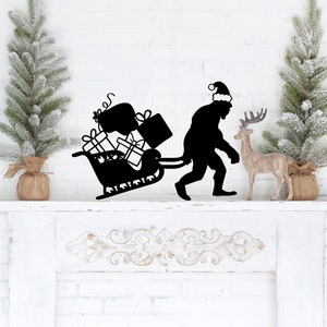 May include: Black metal silhouette of a Bigfoot wearing a Santa hat pulling a sleigh full of presents. The silhouette is on a white mantel with two small, snow-covered Christmas trees on either side.