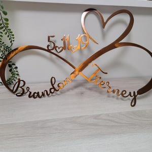 May include: A copper-colored metal infinity sign with a heart and the date 5/11/19. The names "Brandon" and "Tierney" are written in cursive script. The sign is a decorative item.