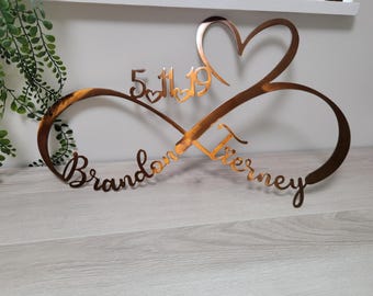 May include: A copper-colored metal infinity sign with a heart and the date 5/11/19. The names "Brandon" and "Tierney" are written in cursive script. The sign is a decorative item.