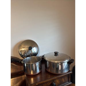 Può includere: Un pentolino in acciaio inossidabile con manico nero e coperchio abbinato, accanto a una pentola più grande in acciaio inossidabile con due manici laterali neri e coperchio. Entrambi i recipienti hanno una base color rame e finitura lucida.