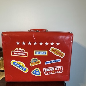 May include: A red vintage suitcase adorned with white star stickers and travel destination labels, including "Paramount," "Broadway," and "Las Vegas." The suitcase has a handle and metal hardware.