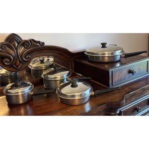 Puede incluir: Un conjunto de cuatro ollas y sartenes de acero inoxidable con tapas a juego y mangos negros. El set incluye varios tamaños de recipientes de cocina, cada uno con un acabado metálico pulido y un diseño sencillo y funcional para cocinar.