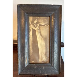 May include: A sepia-toned framed print featuring a classical figure in flowing robes, reaching towards a floral arrangement. The artwork is set within a dark, rectangular wooden frame, adding a vintage aesthetic. The frame has a distressed finish.