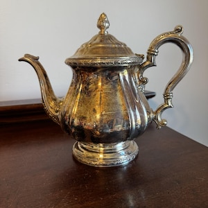 May include: A silver-plated teapot featuring a curved spout, a decorative handle, and a removable lid with a finial. The body of the teapot has a polished finish with visible tarnishing and intricate embossed detailing along the base and rim.