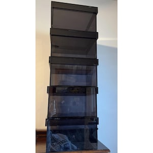 Puede incluir: Una pila de cinco cajas rectangulares transparentes con tinte negro. Cada caja tiene un marco negro y un cuerpo transparente, lo que permite vislumbrar el contenido. Las cajas están dispuestas verticalmente, creando una exhibición.