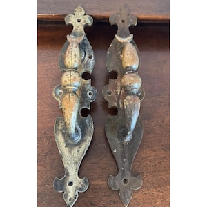 May include: Two antique metal door pulls with ornate designs. Each pull features a decorative top, a central bulbous element, and a stylized handle. The metal has a weathered, aged appearance with shades of brown and gold.