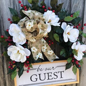 May include: A white magnolia wreath with a burlap bow and red berries. The wreath has a sign that says "be our GUEST".