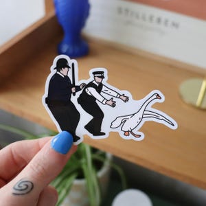 Op de afbeelding: Een sticker met een cartoonillustratie van twee politieagenten in zwarte uniformen die proberen een gans te vangen. De gans is wit met oranje details. De sticker wordt vastgehouden door een hand met blauwe nagellak.