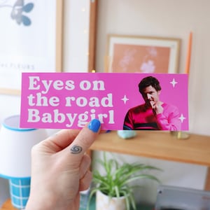May include: A pink bumper sticker with the text "Eyes on the road Babygirl" in white. The sticker features an image of a person in a pink sweater. White star graphics are on the right side of the sticker.
