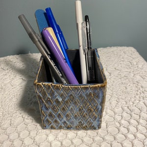 May include: A blue ceramic pencil holder with a textured surface holds a variety of writing utensils, including a black pen, a white pen, a purple pen, a blue pen, a gray pen, and a beige highlighter.