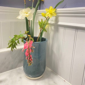 May include: A blue ceramic vase with a white rim, filled with yellow daffodils, white flowers, and pink bleeding heart flowers.