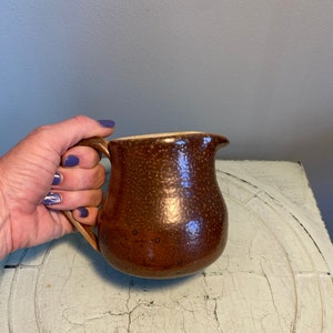 May include: A brown ceramic creamer with a speckled glaze and a rounded shape. The creamer has a handle and a spout.