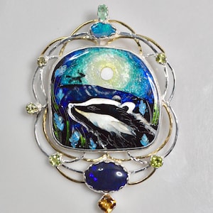 Op de afbeelding: Een zilveren en gouden hanger met een das in een maanverlicht boslandschap. De hanger is omgeven door een decoratief frame met groene en blauwe edelstenen.