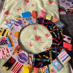 May include: A colorful baby sensory toy with a variety of textures and patterns. The toy features a ring of fabric with different designs, including crayons, planets, and a rocket ship. The ring is attached to a variety of ribbons and fabric scraps in different colors and patterns.