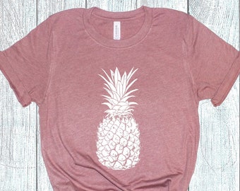Pode incluir: Camiseta rosa empoeirada com um grande desenho de abacaxi branco. O abacaxi está centralizado na camiseta e é muito detalhado. A camiseta tem mangas curtas e gola redonda. O tecido parece macio e ligeiramente desgastado.
