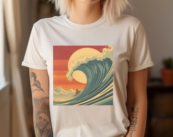 May include: A cream-colored t-shirt featuring a graphic of a large ocean wave with a setting sun in the background. The wave is depicted in shades of blue and white, with a small figure on a surfboard. The shirt is worn by a person.