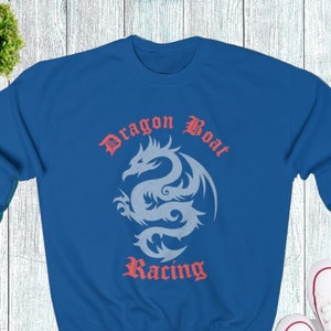Puede incluir: Una sudadera azul real con un diseño de dragón blanco y las palabras "Dragon Boat Racing" en una fuente gótica. Zapatillas de lona blancas con detalles rojos se colocan debajo. Una pequeña planta verde está en la esquina superior izquierda.
