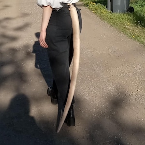 May include: A person wearing black pants and a white blouse with a long, light-colored tail attached to the pants. The person is walking down a dirt road on a sunny day.