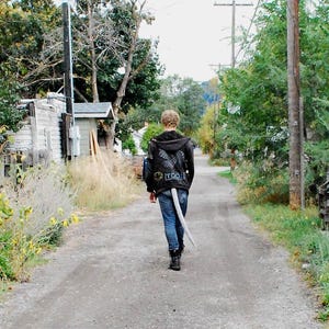 May include: A person walking down a dirt road, wearing a black jacket with a winged design and the word "TOOL" on the back. They are wearing blue jeans and black boots, and have a white tail attached to their jeans.