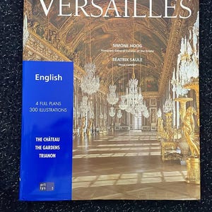 May include: A book titled "Your Visit to Versailles" with a photograph of a grand hall with chandeliers. The book cover has the text "English", "4 FULL PLANS", and "300 ILLUSTRATIONS". The book also lists "THE CHÂTEAU", "THE GARDENS", and "TRIANON".