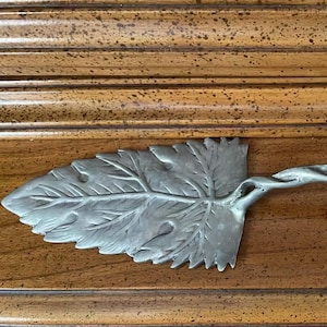 May include: Silver-colored cake server with a leaf-shaped blade and a handle designed to resemble a twisted branch. The server is made of metal and rests on a wooden surface.