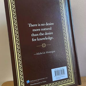 Puede incluir: Un libro de tapa dura con una cubierta marr&oacute;n y un borde decorativo dorado. El libro muestra una cita de Michel de Montaigne: "No hay deseo m&aacute;s natural que el deseo de conocimiento."
