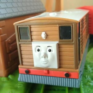 May include: A brown toy train with a smiling face, red accents, and a red cargo car. The train is on a green track with a brown building in the background. The train is a toy.