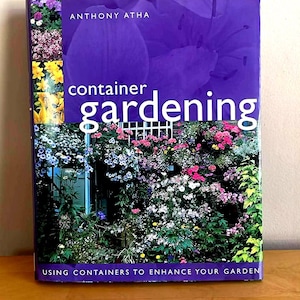 May include: A book cover titled "Container Gardening" by Anthony Atha. The cover features a vibrant image of a garden with colorful flowers and greenery. The text "USING CONTAINERS TO ENHANCE YOUR GARDEN" is also visible.