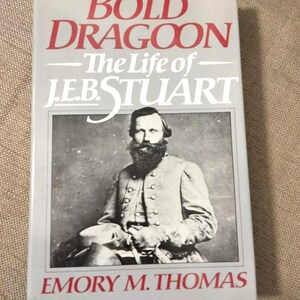 May include: A book titled "Bold Dragoon - The Life of J.E.B. Stuart" by Emory M. Thomas. The cover features a black and white portrait of a man with a long beard in a military uniform. The title is in red, and the author's name is in red.