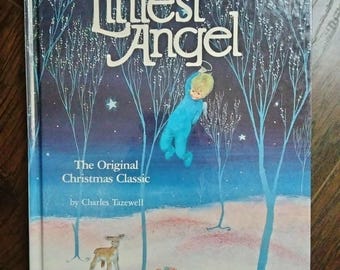 Può includere: Un libro vintage con copertina rigida intitolato "The Littlest Angel" con una copertina blu. L'illustrazione di copertina raffigura un piccolo angelo in abiti blu, appeso a un albero in una scena invernale. Il libro è un classico natalizio di Charles Tazewell.
