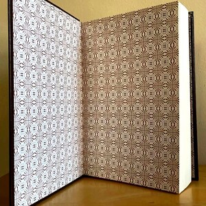 Puede incluir: Un libro abierto con una cubierta marr&oacute;n y p&aacute;ginas blancas. Las cubiertas interiores presentan un patr&oacute;n repetitivo de dise&ntilde;os florales marrones y blancos. El libro se muestra sobre una superficie de madera.