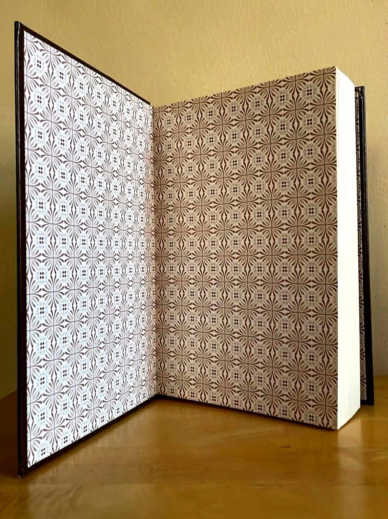 Puede incluir: Un libro abierto con una cubierta marr&oacute;n y p&aacute;ginas blancas. Las cubiertas interiores presentan un patr&oacute;n repetitivo de dise&ntilde;os florales marrones y blancos. El libro se muestra sobre una superficie de madera.