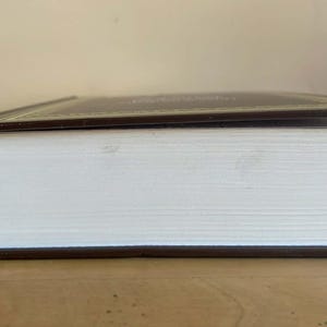 Puede incluir: Un libro de tapa dura con una cubierta marr&oacute;n y bordes dorados. Las p&aacute;ginas son blancas y el libro est&aacute; cerrado, con la cubierta ligeramente entreabierta. El libro est&aacute; sobre una superficie de madera.