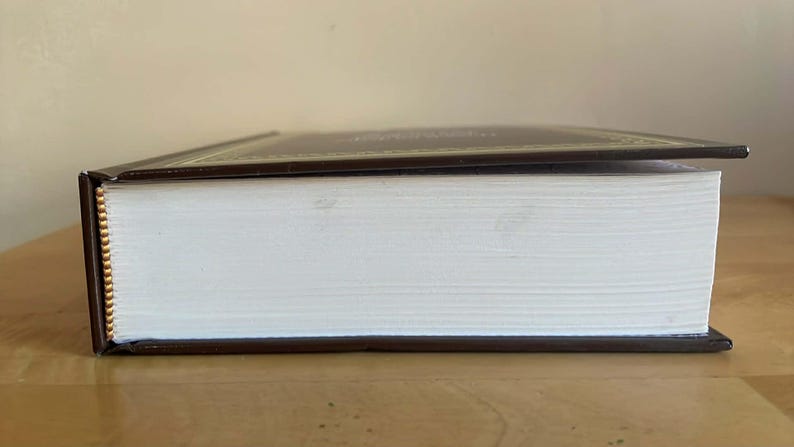 Puede incluir: Un libro de tapa dura con una cubierta marr&oacute;n y bordes dorados. Las p&aacute;ginas son blancas y el libro est&aacute; cerrado, con la cubierta ligeramente entreabierta. El libro est&aacute; sobre una superficie de madera.