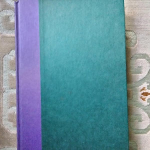 Puede incluir: Un libro de tapa dura con una columna vertebral púrpura y una cubierta verde azulada.