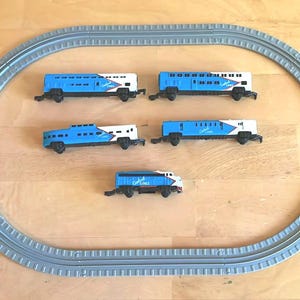 May include: A toy train set featuring five blue and white train cars and a gray oval track. The train cars have the words "Coast Lines" printed on them. The set is arranged on a wooden surface.