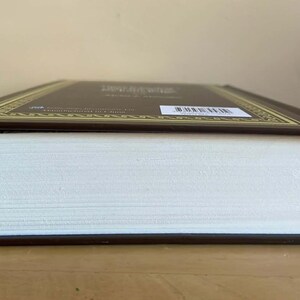 Puede incluir: Un libro de tapa dura con una cubierta marr&oacute;n y un borde decorativo dorado. Las p&aacute;ginas son blancas y el lomo es visible. El libro est&aacute; sobre una superficie de madera.
