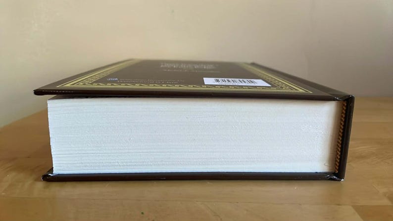 Puede incluir: Un libro de tapa dura con una cubierta marr&oacute;n y un borde decorativo dorado. Las p&aacute;ginas son blancas y el lomo es visible. El libro est&aacute; sobre una superficie de madera.