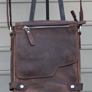 May include: A brown leather crossbody bag with a zipper closure and a flap. The bag has a silver zipper pull and two silver rivets on the bottom.