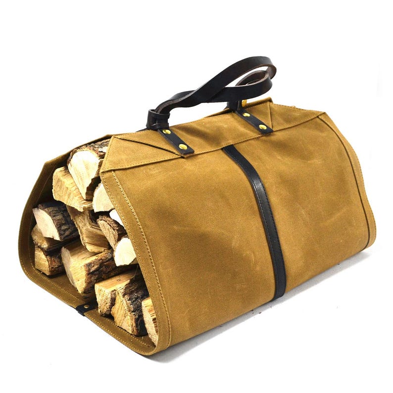May include: A brown canvas log carrier with a black leather handle and accents. The carrier is filled with cut firewood. The handle is secured with brass rivets. A black leather strap runs down the front of the carrier.