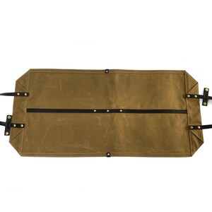 May include: A brown waxed canvas tool roll with black leather straps and brass rivets. The roll is rectangular with a flat bottom and top, and has two black leather straps running across the middle. The handles are black leather.